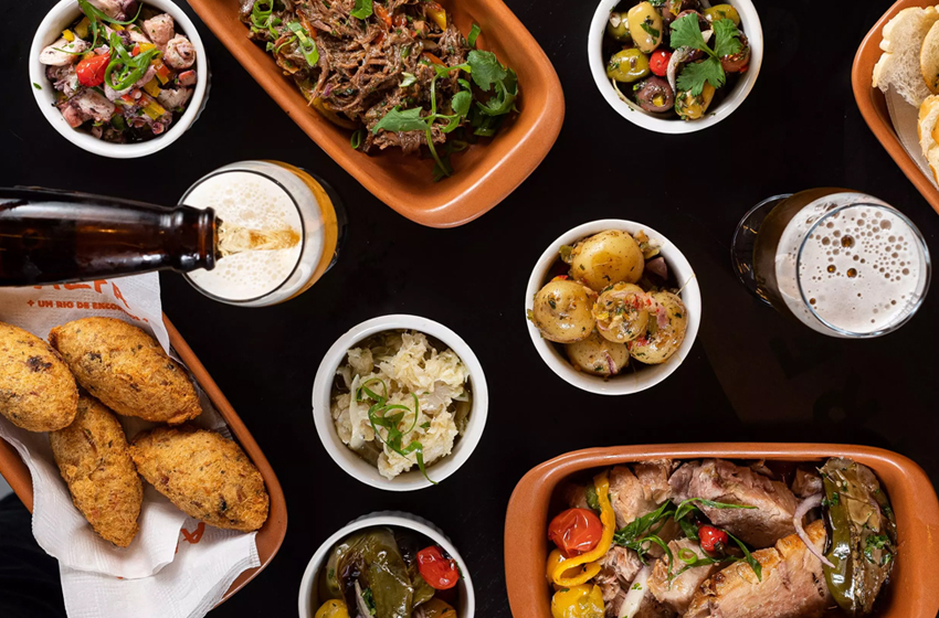 cerejas, carne de desfiada, e demias prats de comidas brasileiras dipostas em uma mesa de fundo escuro
