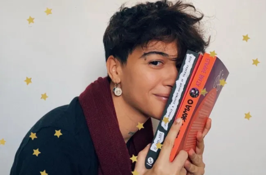 escritora, usando cabelos curtos, brincos, blusa azul e cachecol vermelho segurando seus dois livros publicados