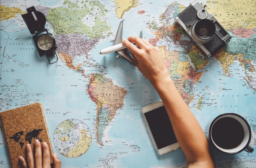 mão humana segurando um avião de brinquedo sobre um mapa mundi, e artigos relacionados a viagem e exploração espalhados sobre a mesa