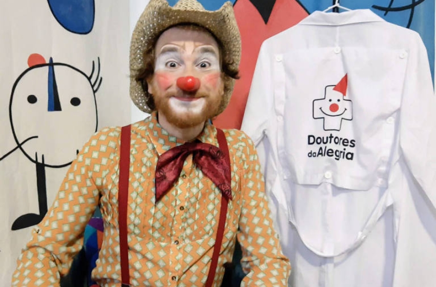 integrante dos doutores da alegria posando ao lado de jaleco com a logo do projeto