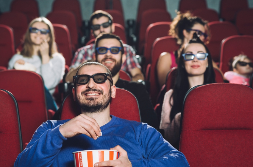 homem usando blusa azul e assistindo um filme em 3d na sala de cinema com pessoas ao fundo
