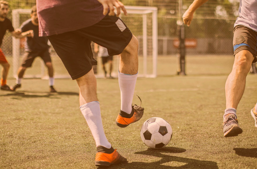 A foto mostra um homem de chuteira laranja, meiões brancos, shorts pretos e blusa vinho. Ele passa o pé por cima de uma bola de futebol, com quadrados pretos e brancos. O marcando, está um homem de tênis acinzentados, short cinza com detalhe em azul e uma camisa em azul escuro. Eles estão em uma quadra de futebol, com a grama verde e o gol ao fundo da imagem.