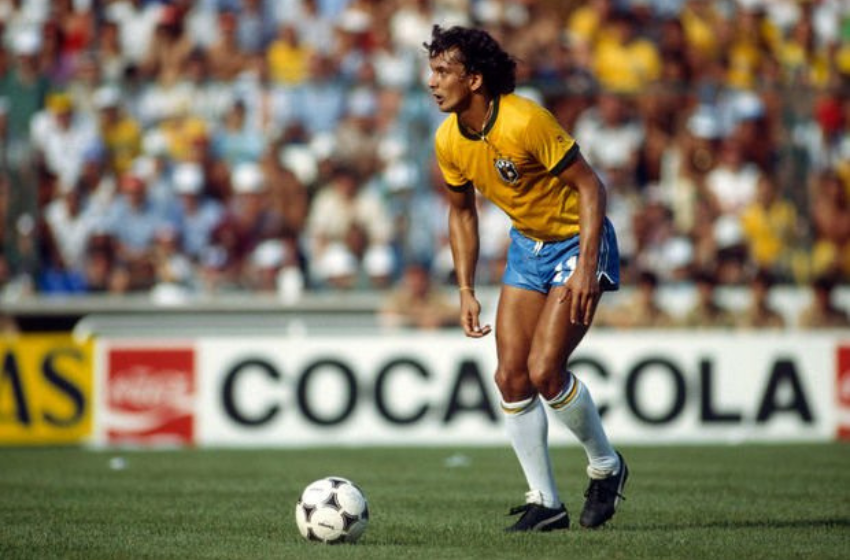 Na foto está um homem branco, com uma camisa amarela, shorts azuis e meiões brancos. Ele esta um pouco abaixado, indo em direção a uma bola de futebol branca, com detalhes triangulares pretos. Ele está em um gramado verde com uma enorme torcida atrás.
