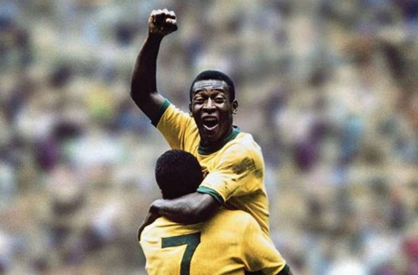 Na foto está Pelé, um jogador negro que comemora o seu gol com o punho fechado e o braço levantado. Ele está vestido uma camisa amarela e está sendo segurado por Jairzinho, um outro jogador negro que veste uma camisa amarela com o número 7 escrito em verde nas costas.