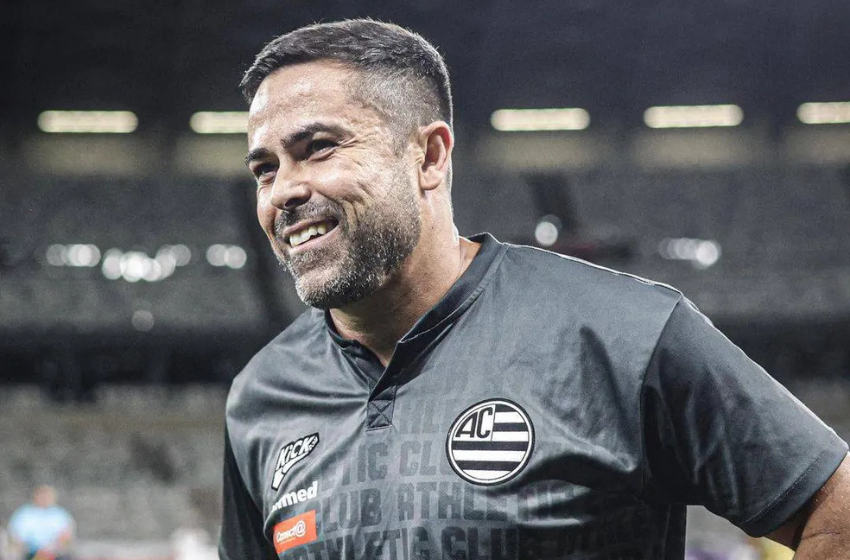 Na foto está Cícero Junior, um homem branco de barba pequena e cabelos pouco grisalhos nas laterais. Ele veste uma camisa cinza chumbo, com o símbolo do Athletic e as escritas Athletic Club em alto relevo. Em seu fundo estão as arquibancadas do Mineirão com suas cadeiras brancas.