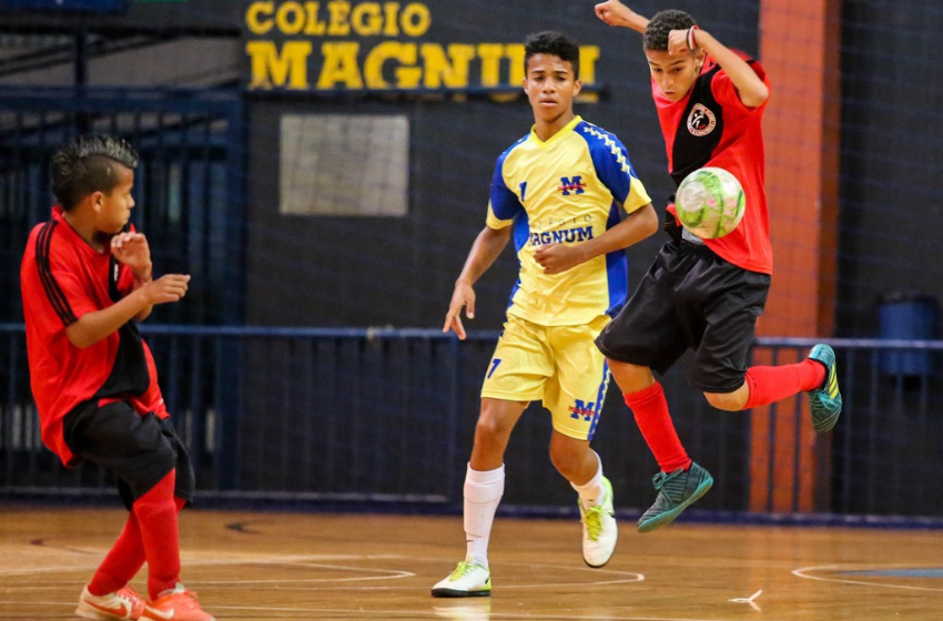 A foto mostra 3 garotos. 2 deles estão com uma blusa vermelha, com uma listra horizontal preta, shorts pretos e meiões vermelhos. O outro menino está com um uniforme amarelo, com detalhes azuis, shorts também amarelos com listras azuis e meiões brancos. Eles disputam os Jogos Escolares de Belo Horizonte, na modalidade Futsal.