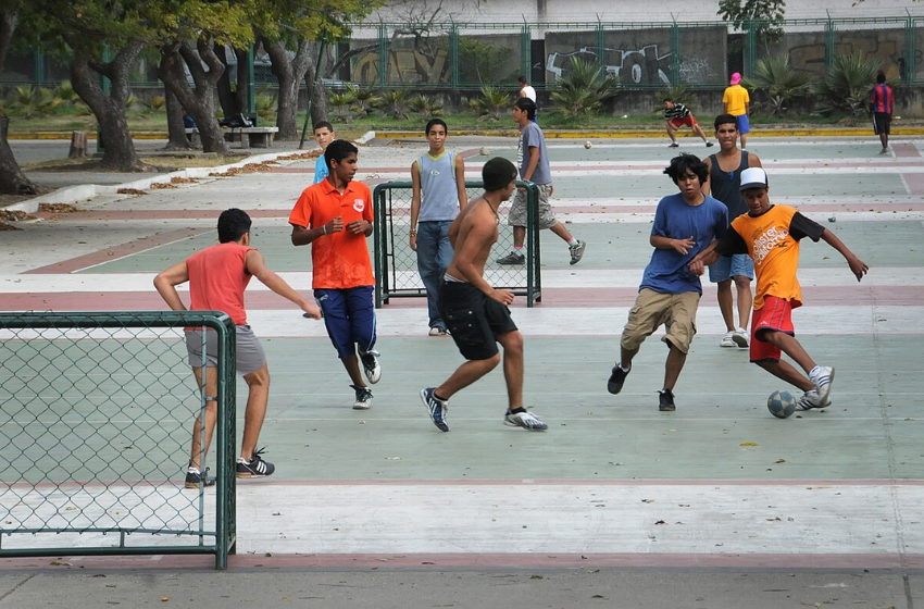 A foto mostra diversos adolescentes. Um deles, posicionado ao centro da imagem, está sem camisa. Os demais estão com suas camisas e bermudas longas. Todos eles estão em uma quadra de cimento, se divertindo com uma pequena bola azul e branca. Eles estão jogando Futebol de Rua, esporte semelhante ao Futsal.
