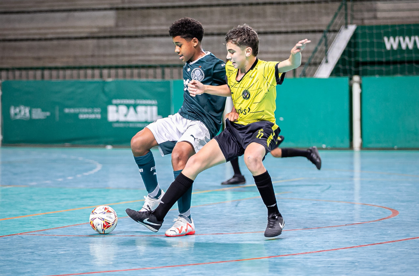 A imagem mostra um menino de camisa verde, shorts brancos e meiões verdes, disputando uma bola com outro garoto, esse de blusa amarela, shorts e meiões pretos. Os jovens estão em um campeonato de futsal sub-12.