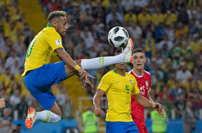 A foto mostra Neymar, na copa de 2018, dominando uma bola no ar, com a perna esticada na horizontal. Ao fundo, observando o lance, está o volante camisa 5, Casemiro. Ambos estão com o uniforme da seleção brasileira, uma camisa amarela, shorts azuis e meiões brancos. Eles se encontram em um estádio de futebol, que tem em sua torcida uma maioria de uniformes amarelos.